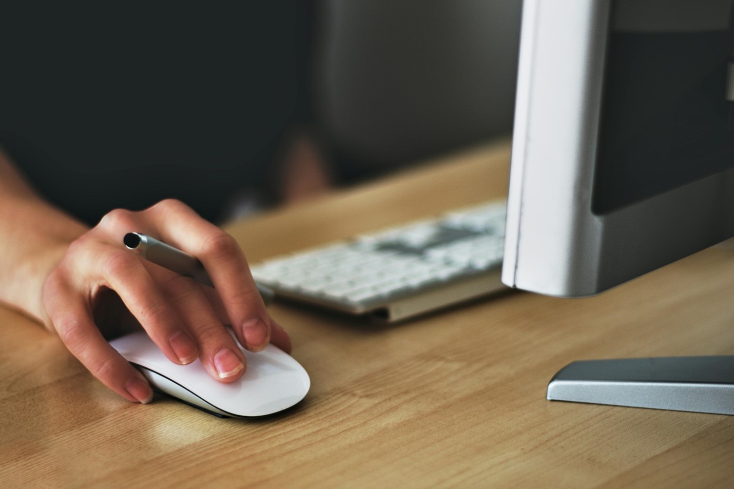 A person using a mouse with a pen in their hand next to a keyboard and monitor.